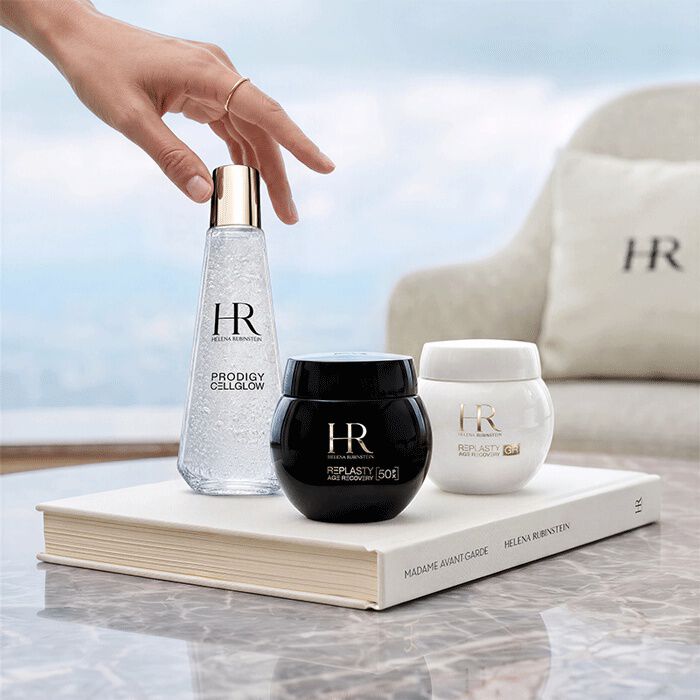 Tre prodotti di skincare Helena Rubinstein (Prodigy Cellglow e Re-Plasty) disposti su un libro bianco davanti a una finestra con vista sul paesaggio.​