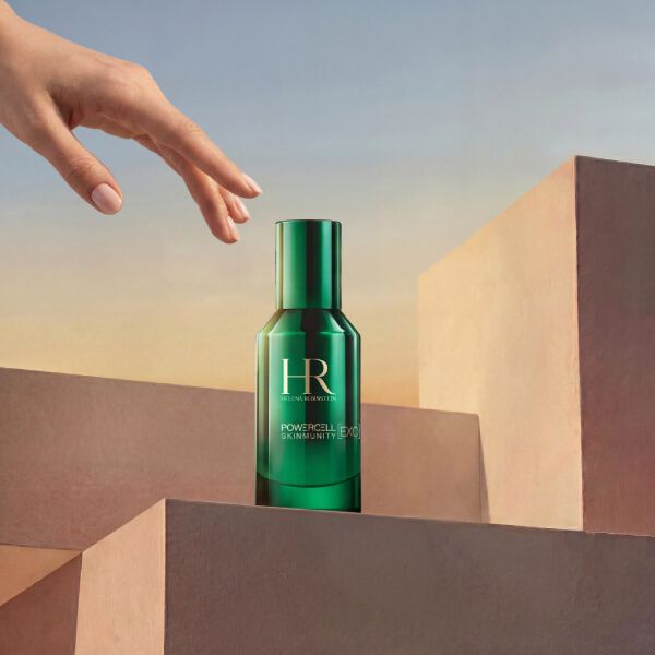 Foto del prodotto Siero Powercell Skinmunity Exo di Helena Rubinstein in un flacone di vetro verde lucido. Il flacone poggia sulla sommita di una pila di podi geometrici rosa, con una mano elegante che si avvicina dall'alto, su uno sfondo di cielo dai toni caldi al tramonto.​