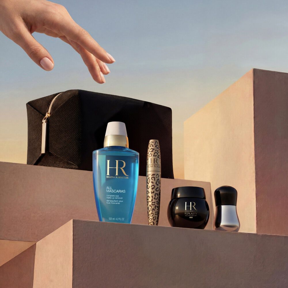 Una mano elegante si avvicina a una selezione di prodotti Helena Rubinstein disposti su blocchi di pietra minimalisti color beige sabbia. Il set e presentato su uno sfondo di cielo dorato al tramonto e include una pochette da viaggio nera, lo struccante occhi blu 'All Mascaras', un mascara Lash Queen Feline Blacks a stampa leopardata, un piccolo vasetto di crema Re-Plasty nero e un applicatore argentato.​