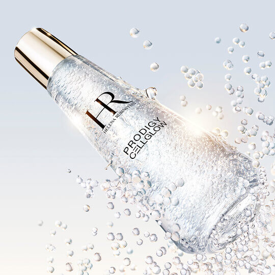 Flacone di siero Helena Rubinstein Prodigy Cellglow riempito di bolle, inclinato e circondato da bolle scintillanti in sospensione, su uno sfondo chiaro e luminoso.