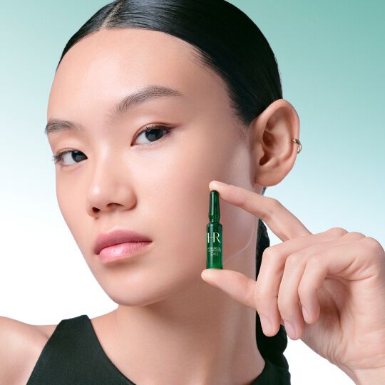 Primo piano del volto di una donna che tiene delicatamente tra le dita un'fiala verde Powercell [EXO] Skinmunity di Helena Rubinstein, su uno sfondo sfumato verde e bianco.​