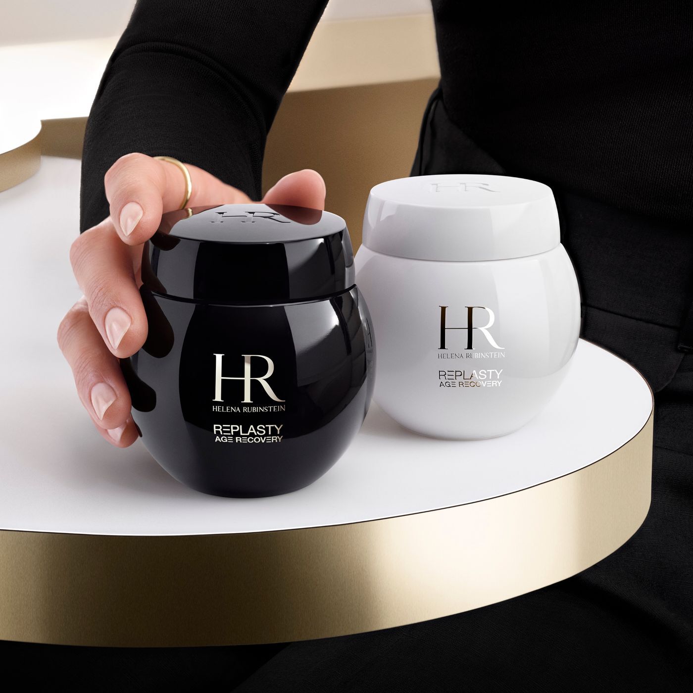 Una mano afferra un vasetto nero di crema Helena Rubinstein Re-Plasty Age Recovery, con un vasetto bianco abbinato accanto, su una piattaforma dorata.