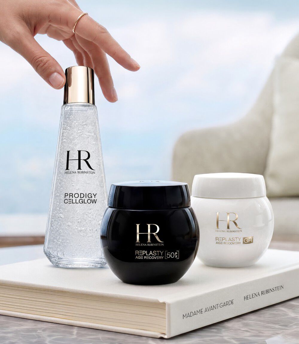 Tre prodotti di skincare Helena Rubinstein (Prodigy Cellglow e Re-Plasty) disposti su un libro bianco davanti a una finestra con vista sul paesaggio.​