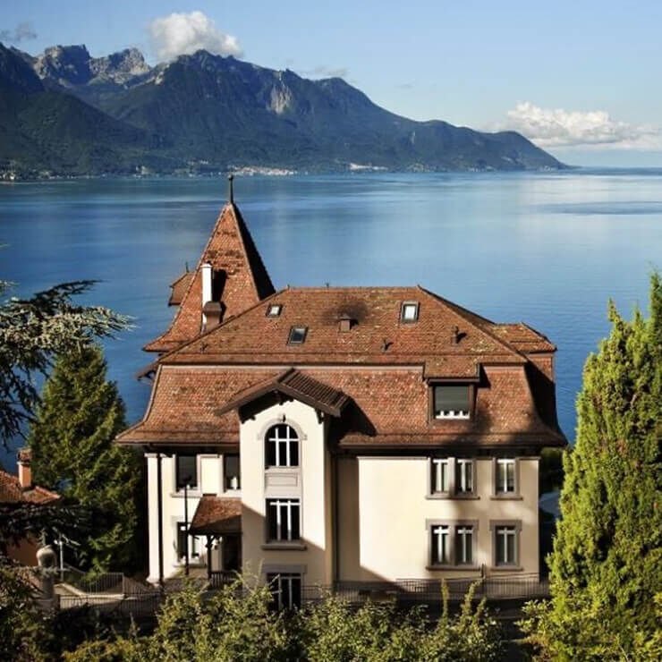 Una grande casa in stile tradizionale con tetto in tegole rosse e pareti chiare, situata sulle rive di un grande lago blu profondo, circondata da montagne. Il cielo e sereno.