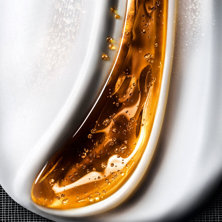 Primo piano di una texture cosmetica fluida, che mescola onde di gel dorato scintillante con bolle su uno sfondo bianco cremoso, evocando una ricca cura della pelle.
