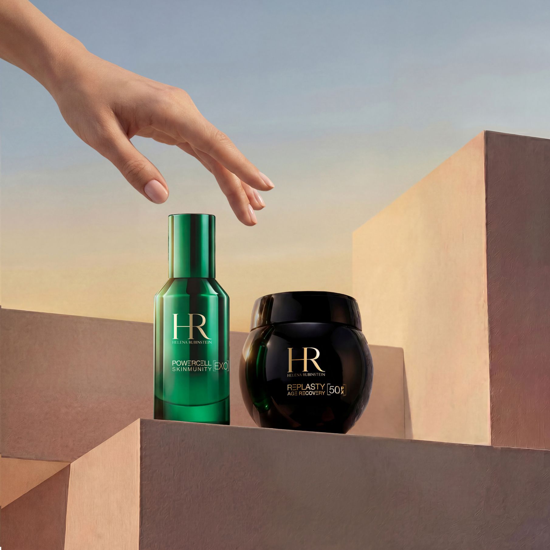 Una mano elegante che raggiunge il Duo da Viaggio Replasty Night – Powercell Essence di Helena Rubinstein, composto da un flacone verde di Powercell Skinmunity Essence e un vasetto di Re Plasty Age Recovery Night Cream, disposti su blocchi geometrici sabbiati su uno sfondo di cielo al tramonto.​