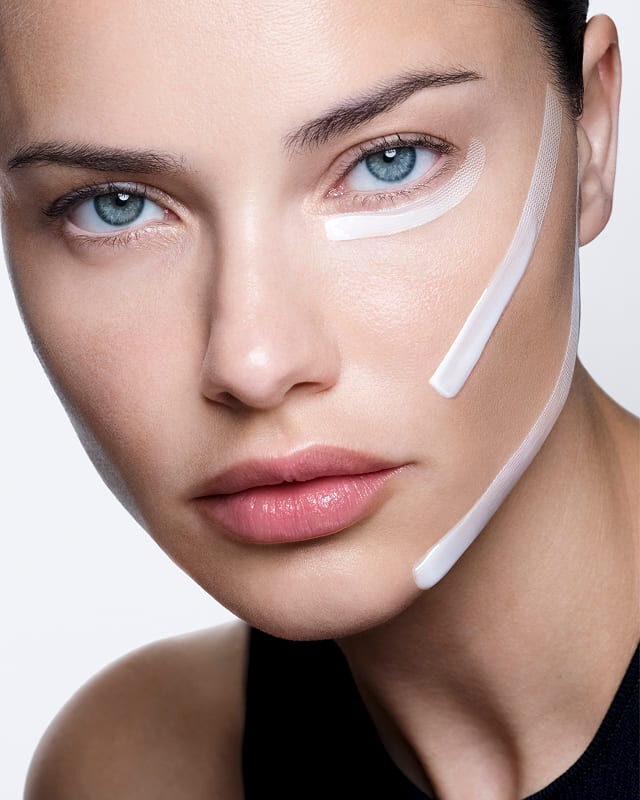 Adriana Lima con crema bianca e strisce applicate sul viso, che dimostrano un effetto lifting visibile.