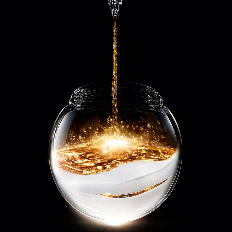 Liquido dorato scintillante si versa in un vaso di vetro sferico, poggiato su una base bianca testurizzata, creando un contrasto luminoso su sfondo nero.