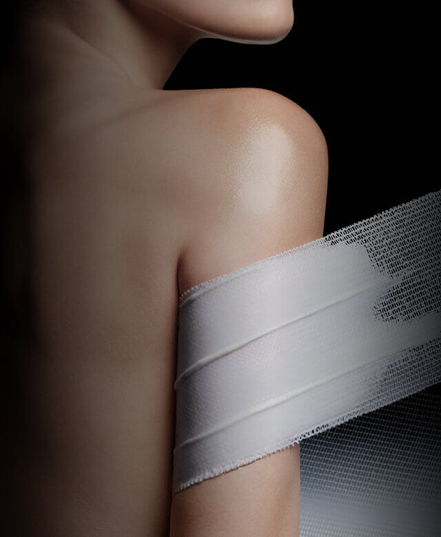 Spalla e braccio nudi parzialmente coperti da una fascia di rete bianca, che suggerisce cura o trattamento della pelle, su uno sfondo scuro.