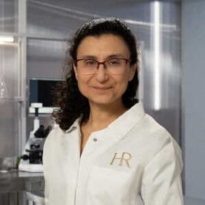 A scientist in a lab coat with 'HR' embroidered, standing in a modern laboratory setting, smiling at the camera.