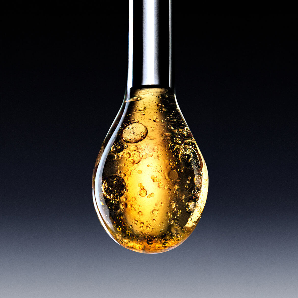 Una grossa goccia di liquido dorato scintillante, come un siero o un olio, che sta per cadere da un applicatore, su uno sfondo scuro sfumato.
