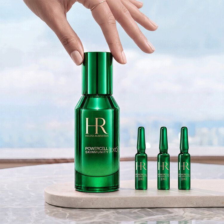 Una mano che tocca il flacone verde del siero Powercell [EXO] Skinmunity di Helena Rubinstein, posizionato accanto a tre fiale [EXO] Shots su un supporto di marmo con vista su un paesaggio luminoso.​