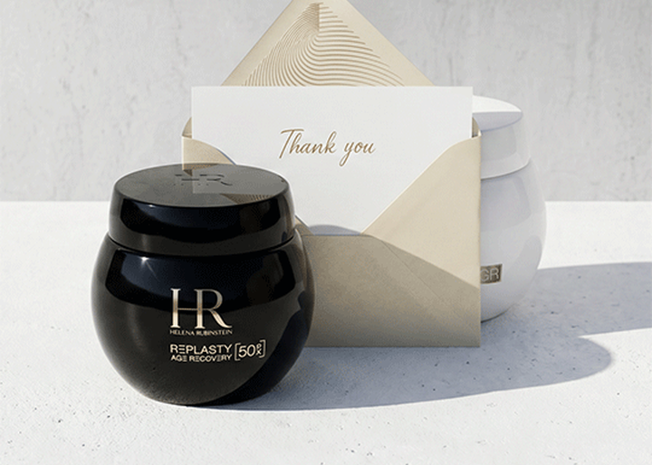 Un barattolo nero di crema Helena Rubinstein Replasty Age Recovery e una busta con un biglietto "Thank you" poggiati su una superficie bianca, con un barattolo bianco sullo sfondo.