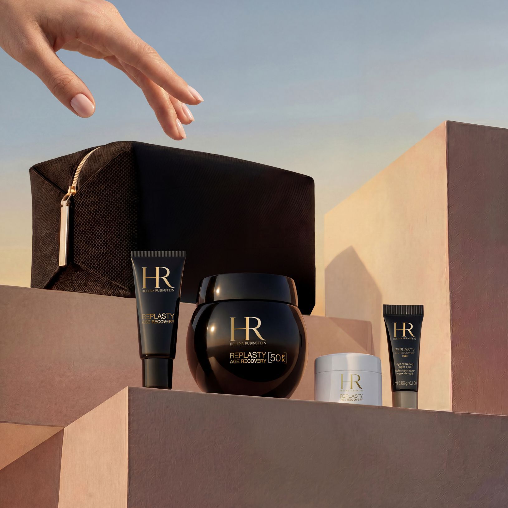 Una mano elegante si avvicina a un cofanetto regalo Helena Rubinstein, disposto su blocchi di pietra sabbiati color beige su uno sfondo di cielo al tramonto, che include una pochette da viaggio in edizione limitata nera e la gamma Replasty Age Recovery: Eye Repairing Night Care, Day Cream, Night 50PX e Body Repairing Care.​